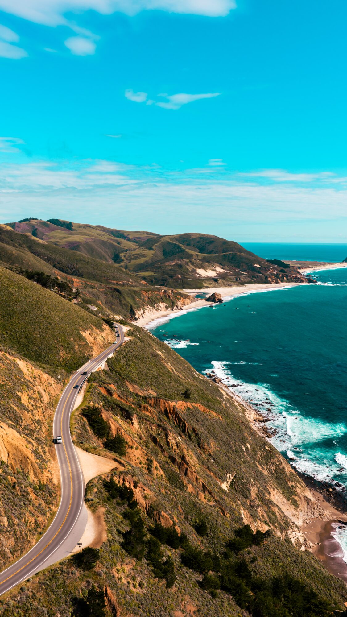 Coastal road winding along cliffs with ocean waves crashing below, hills in the background, under a blue sky.
