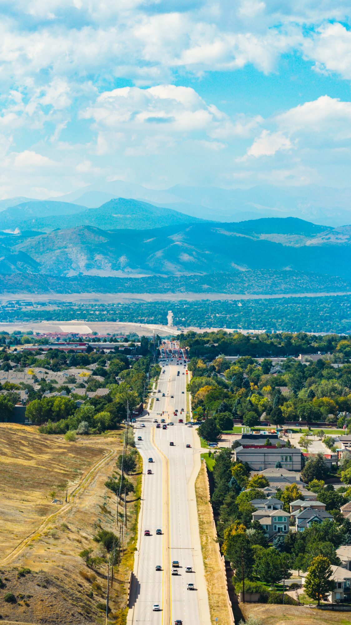 Aerial view of a straight road with cars, surrounded by trees and buildings, leading toward mountains in the distance.