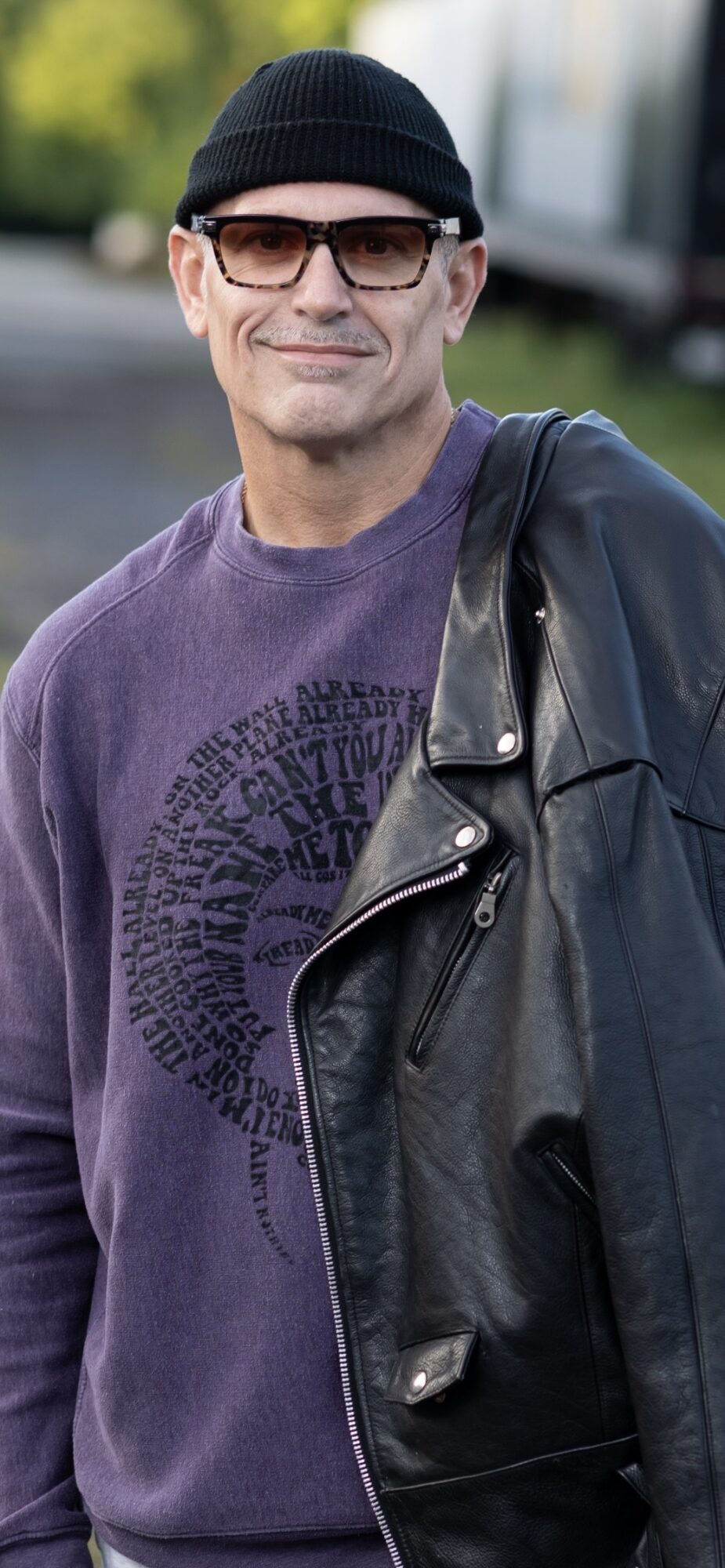 Man wearing glasses, black beanie, purple sweatshirt, and black leather jacket outdoors, smiling.