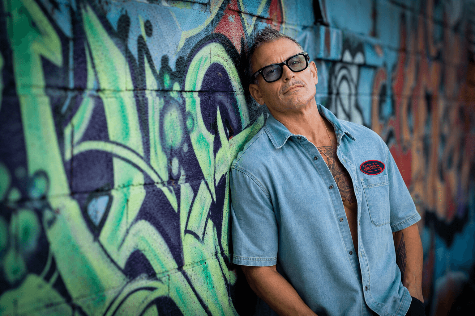 Man wearing sunglasses and a blue shirt stands against a colorful graffiti wall.