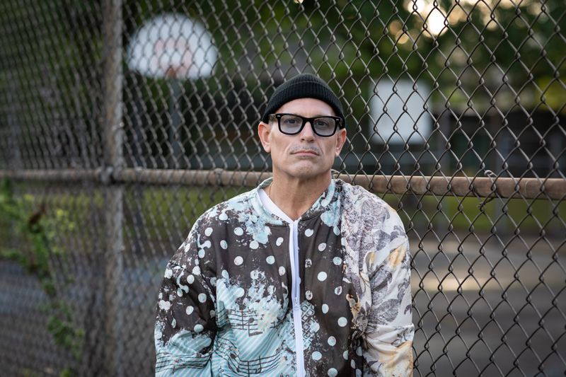 Man wearing glasses and a patterned jacket stands outdoors near a chain-link fence.