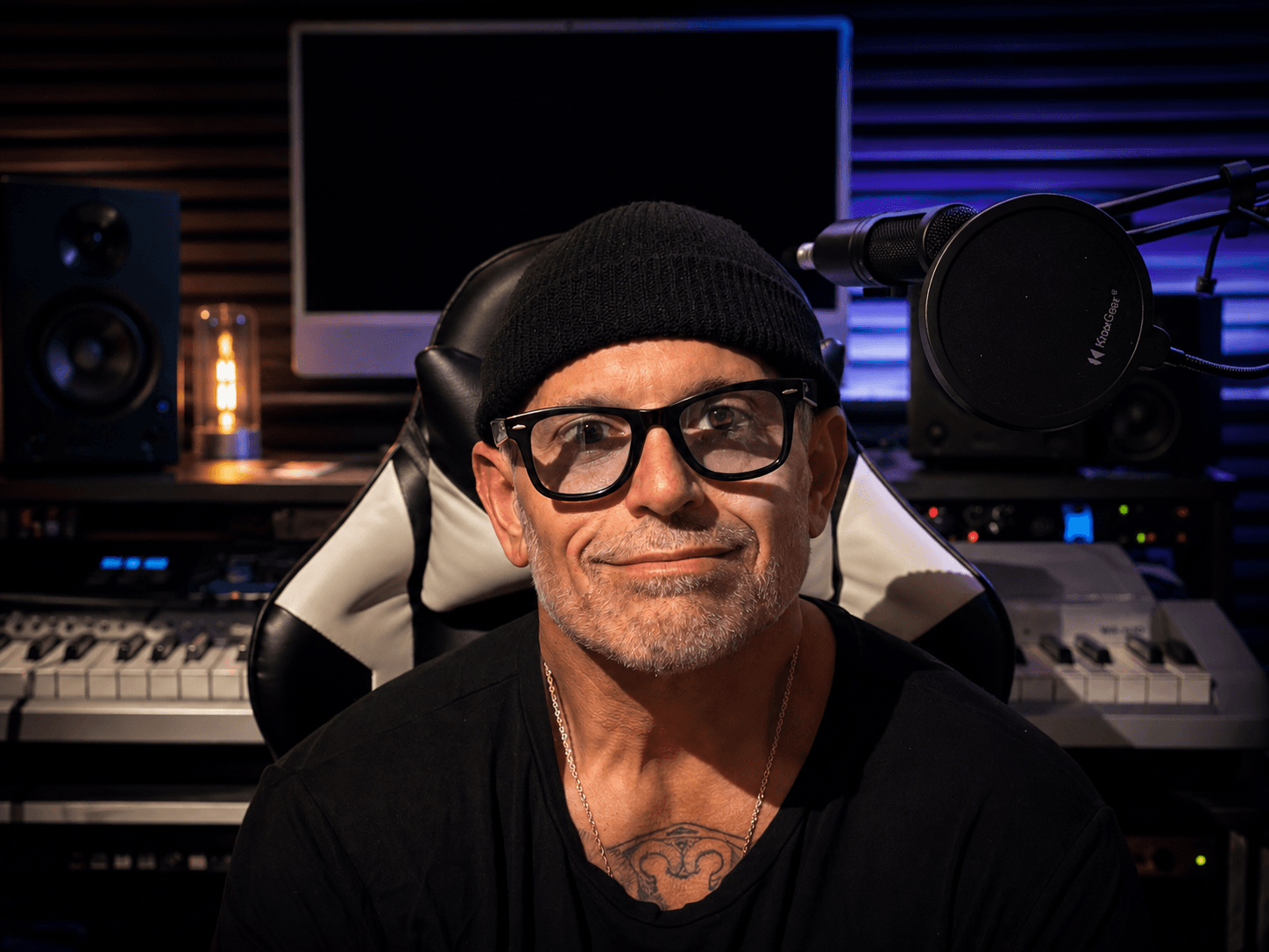 Man wearing glasses and a beanie, sitting in a music studio with equipment behind him.