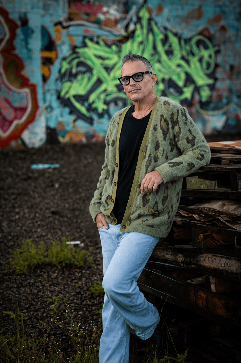 Man with glasses in a green patterned jacket and black shirt leans against a wooden structure in front of graffiti wall