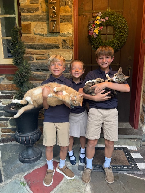 Three children stand outside a house holding cats, smiling, with a wreath on the door and house number 2377 visible.