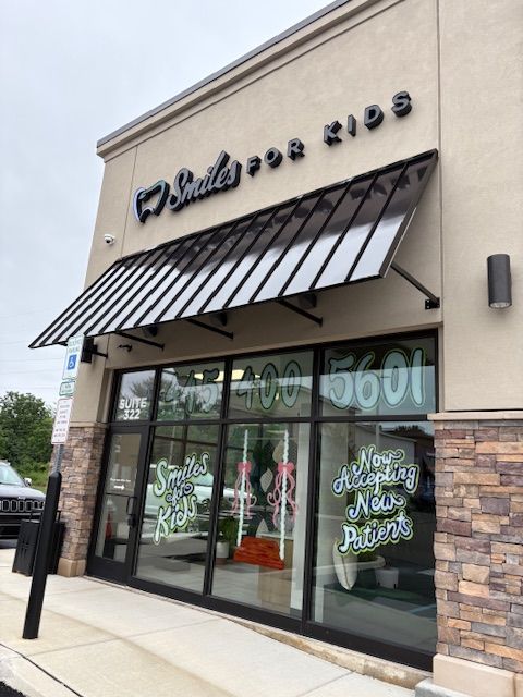 Storefront with glass doors and windows, black awning, and signage, with sidewalk and trees outside.