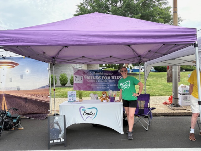 A woman stands behind a table under a purple canopy at an outdoor event, with a banner and promotional materials.