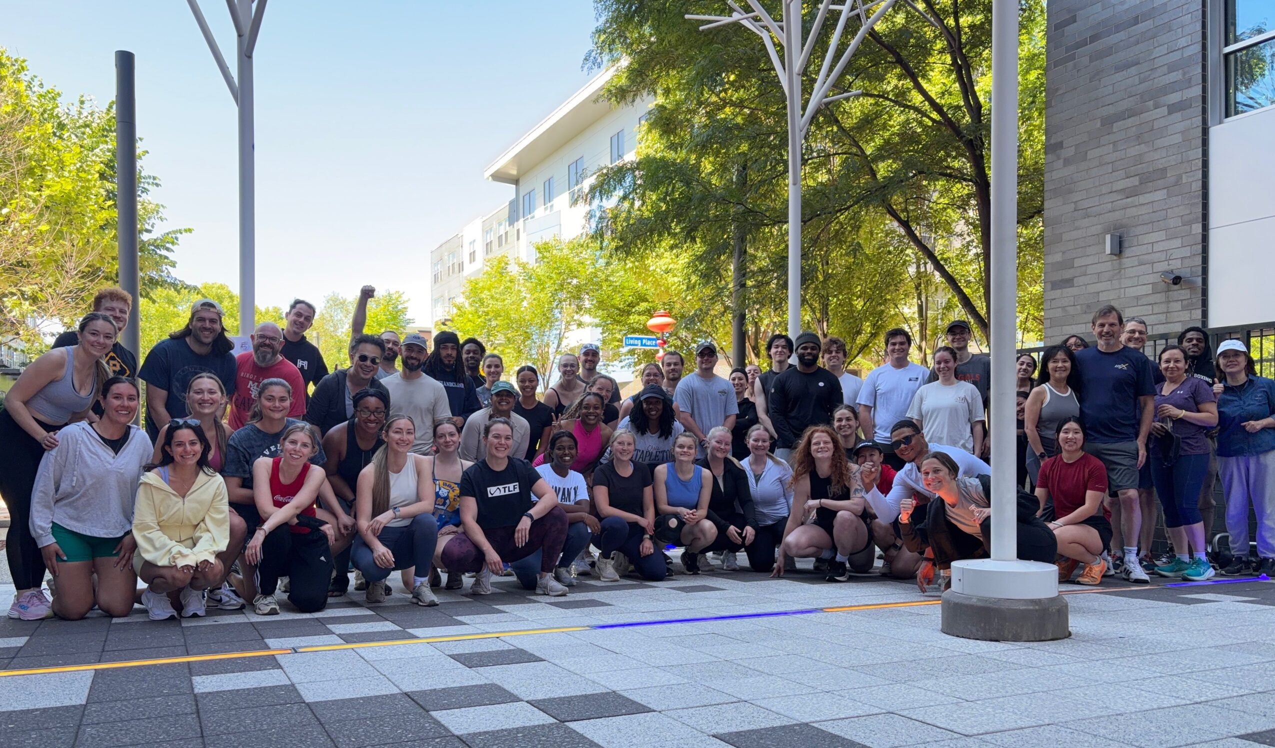 Group of people outdoors on a sunny day, standing and sitting on a paved area with trees and modern buildings in the background.