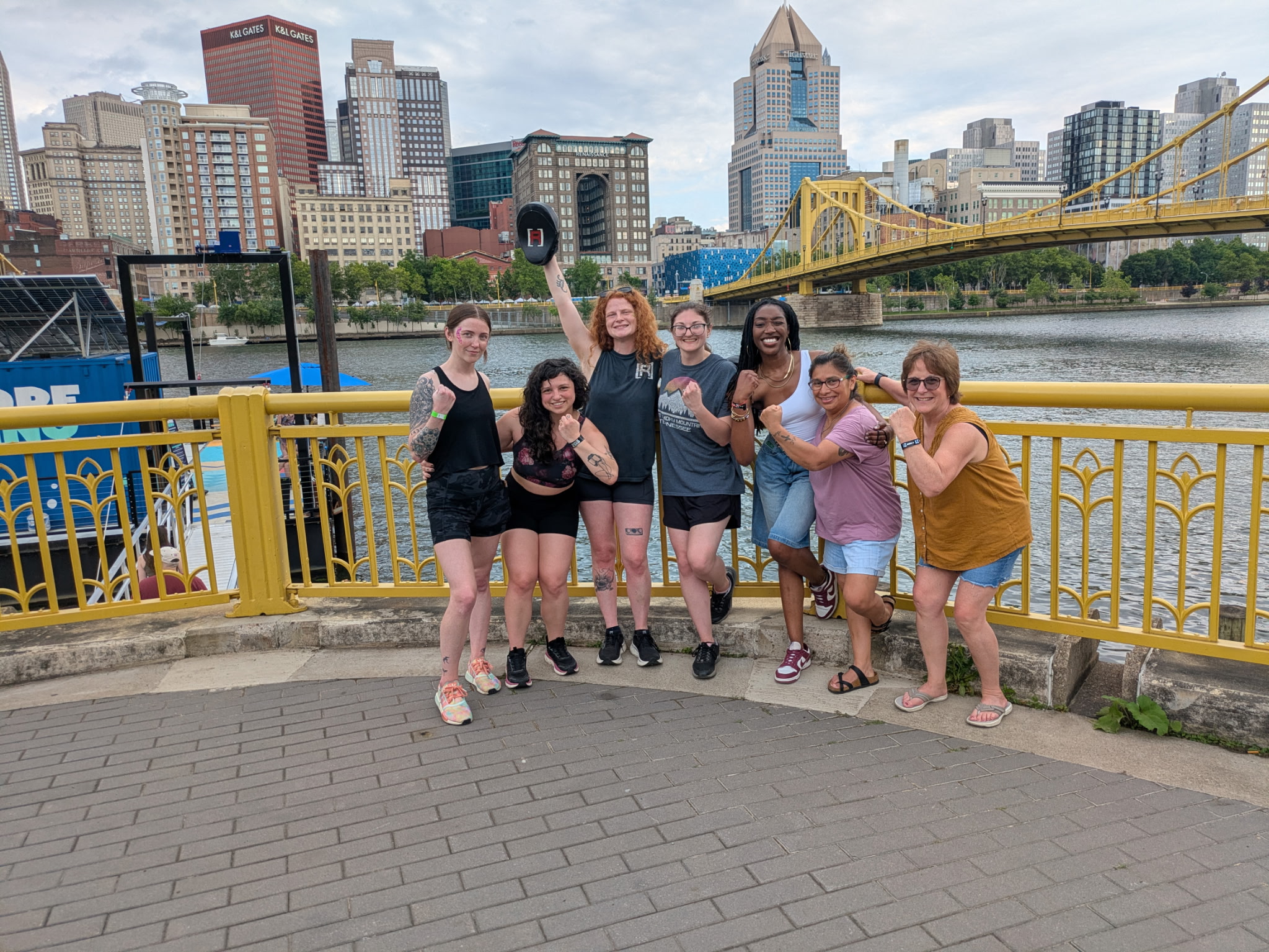 Group of seven people standing by a yellow railing with city skyline and bridge in background, smiling and waving.