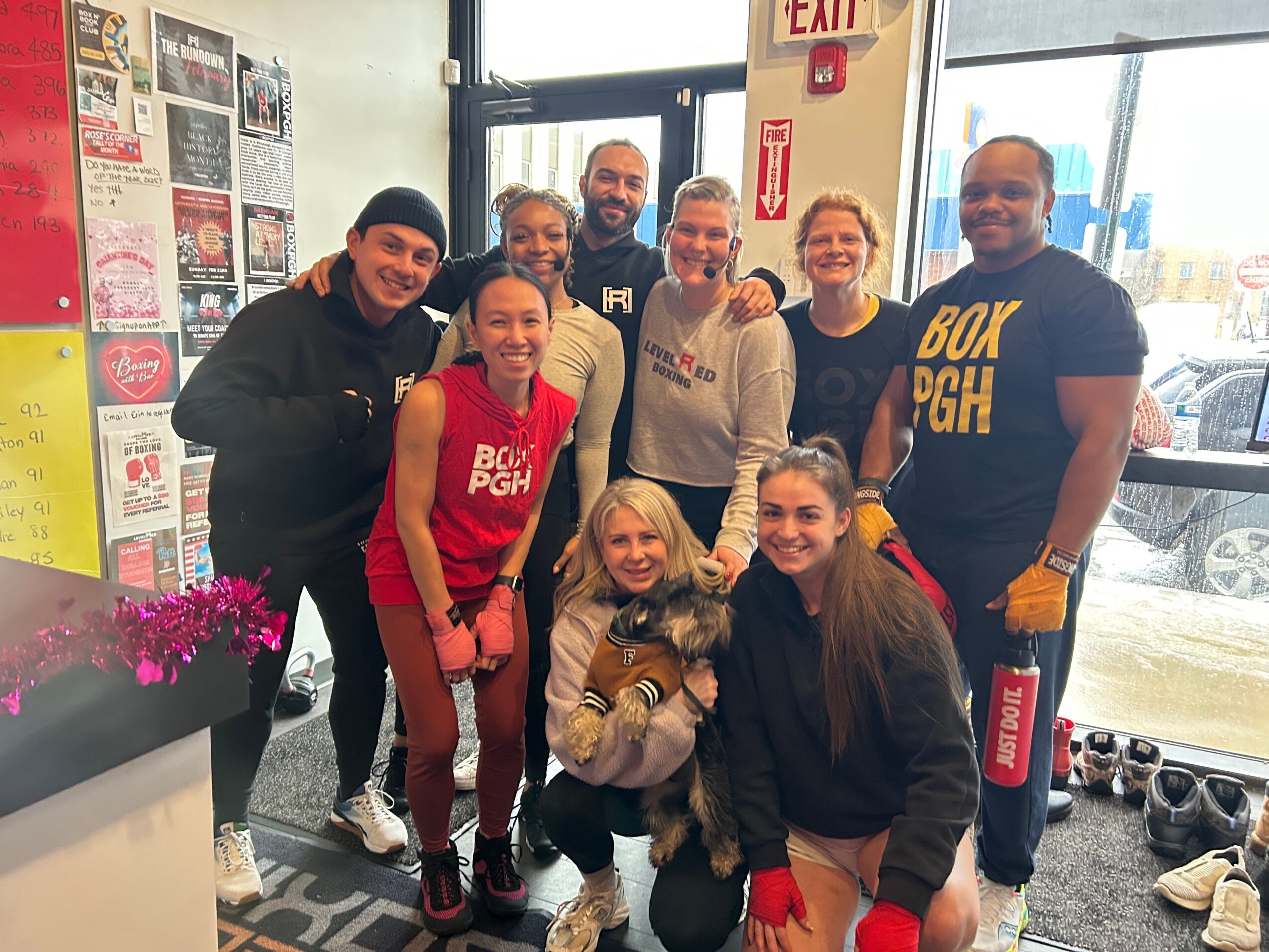 Group of nine people, some wearing red shirts, standing inside a store near a window, with shoes on the floor and posters on the wall.