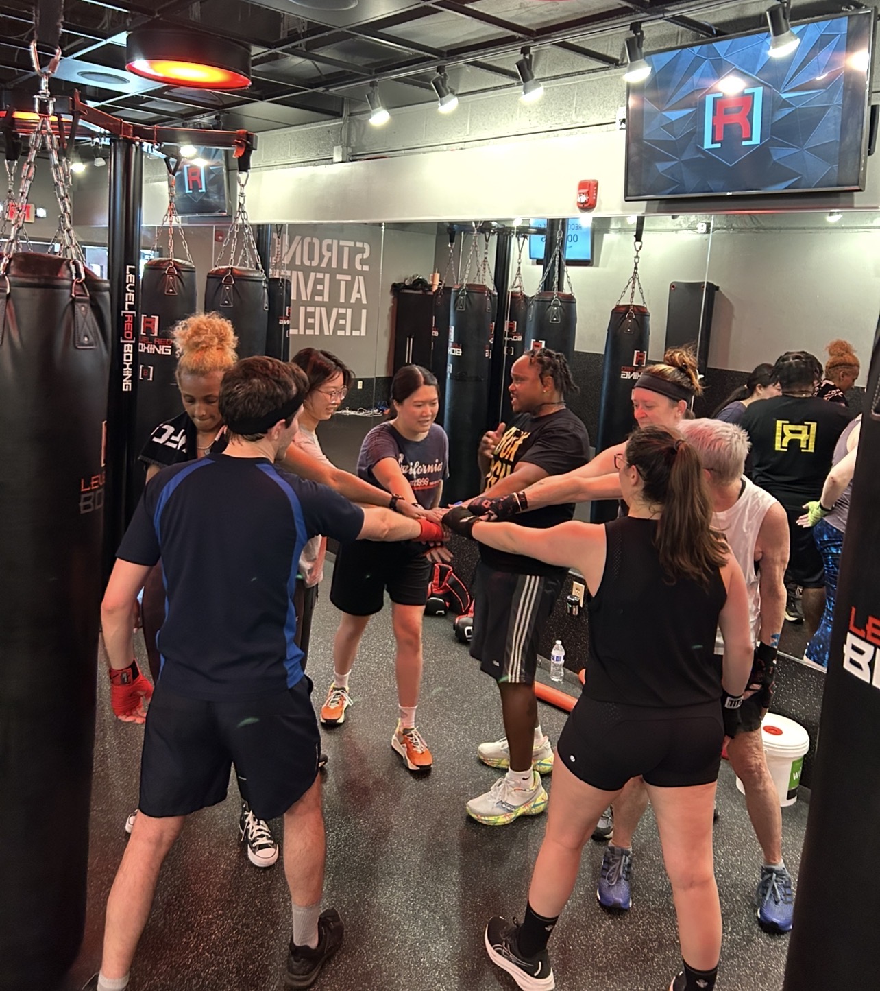 Group of people in a gym, holding hands in a circle, with punching bags and a TV screen in the background.