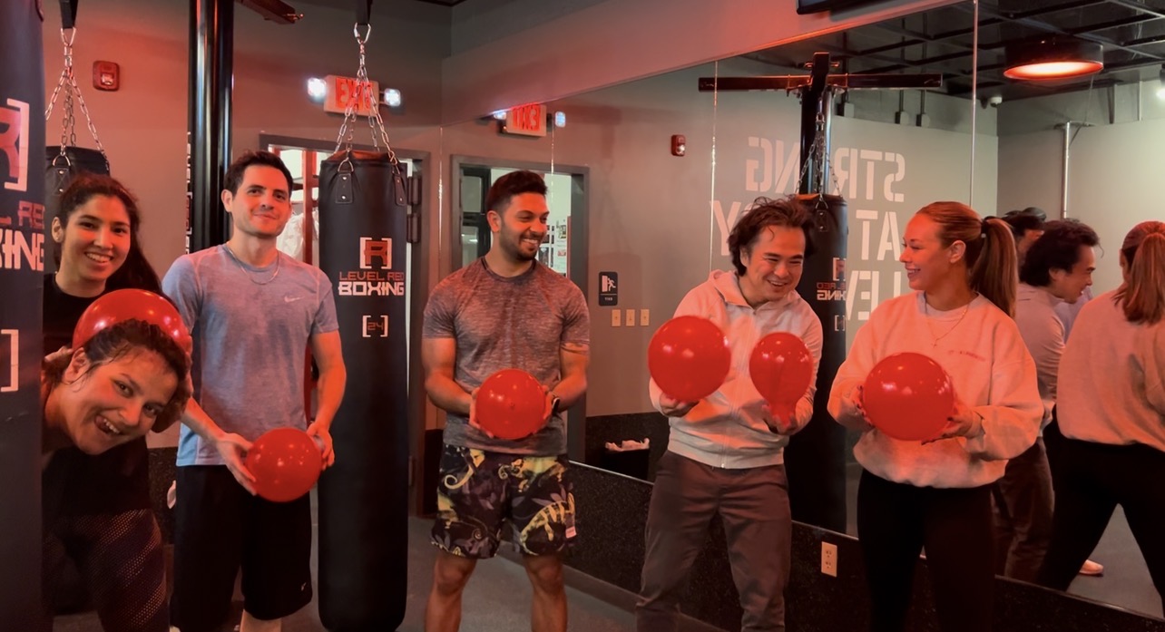 Group of people holding red balloons in a gym with punching bags and exercise equipment.