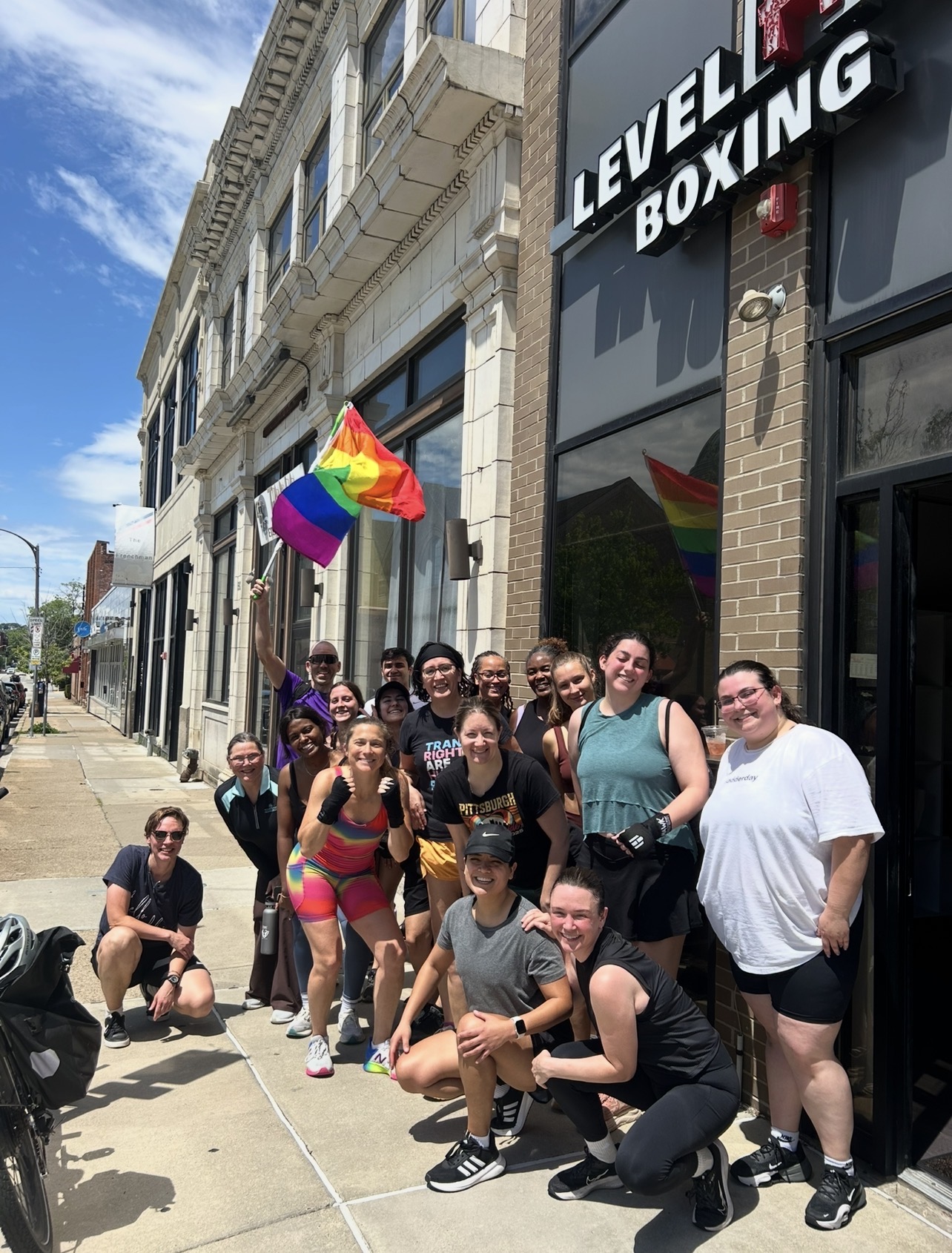 Group of people standing outside a boxing gym with a rainbow flag, smiling and posing for a photo.