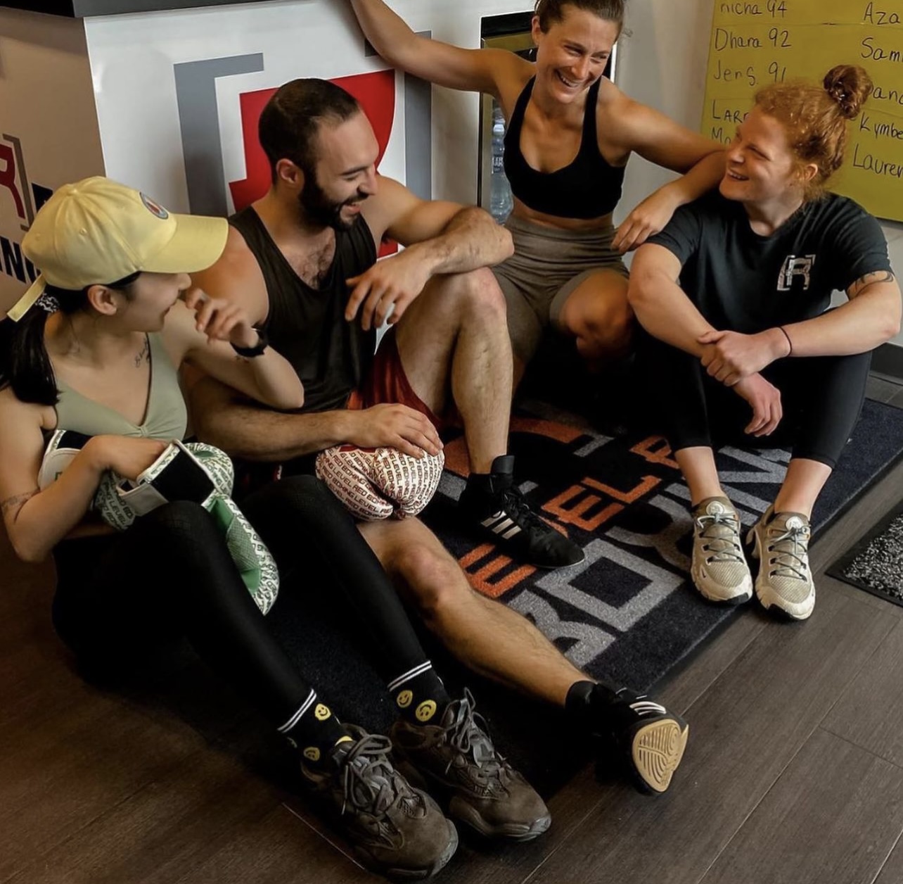 Four people sitting on a floor, smiling and talking, with a yellow board in the background.