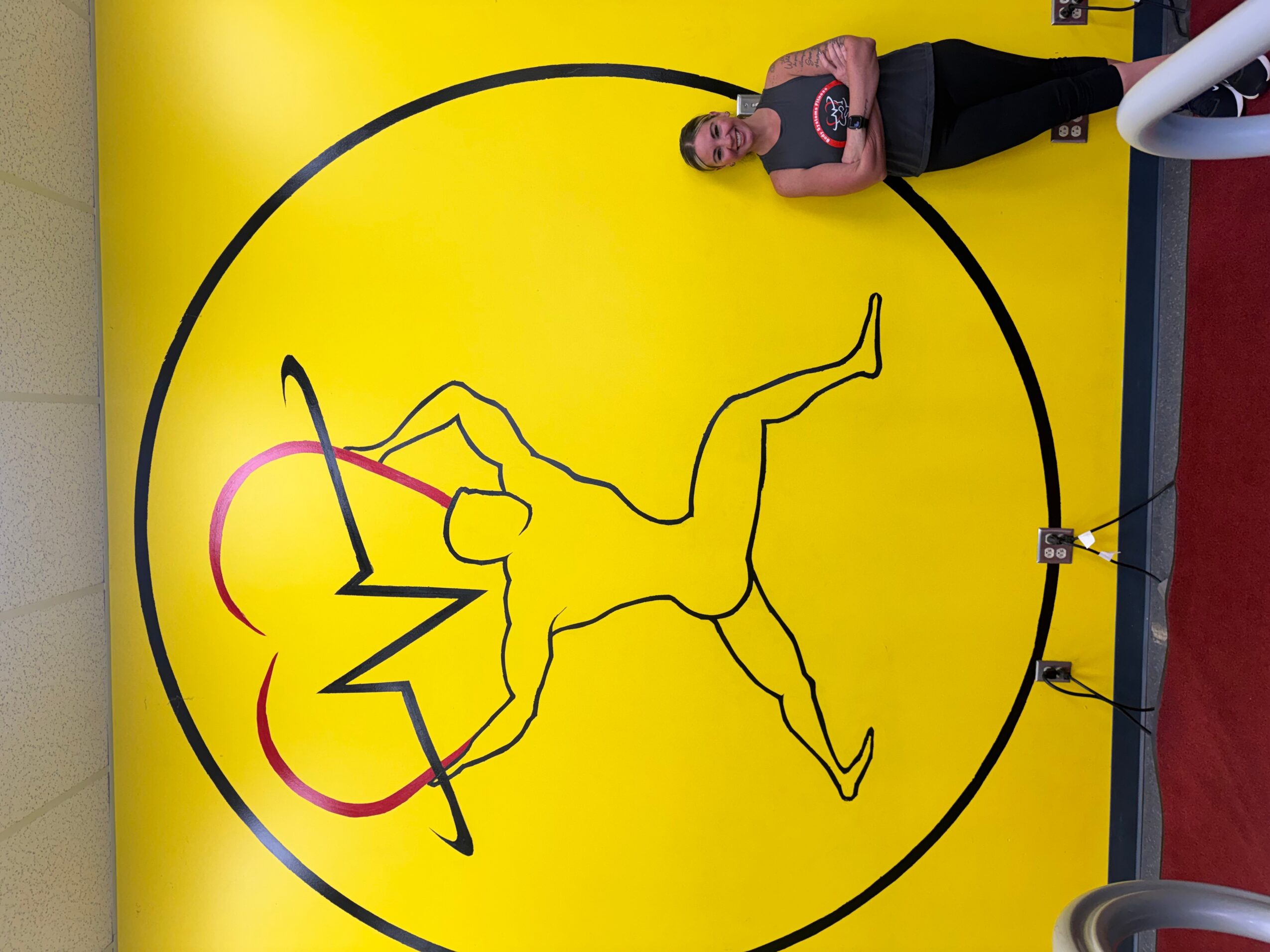 Person standing next to a yellow wall with a black line drawing of a person inside a circle, holding a barbell with a heart rate line.