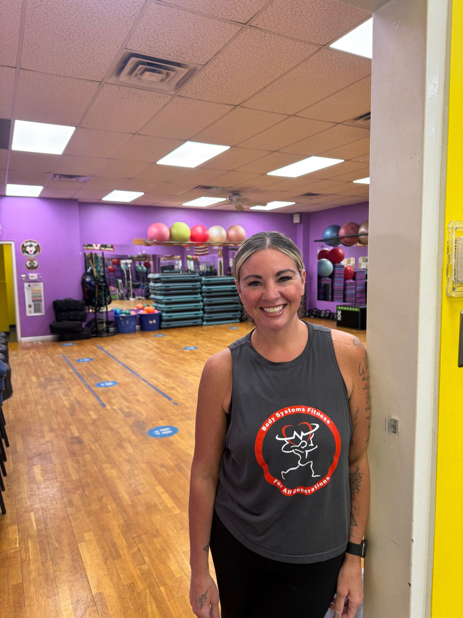 Woman standing in a gym with purple walls, wooden floor, and exercise equipment in the background.