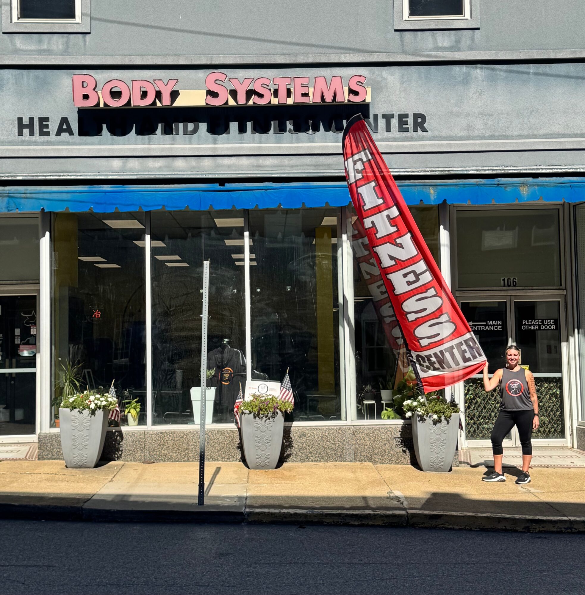 Person standing outside a storefront with large windows, potted plants, and a red vertical banner, in front of a building labeled 'BODY SYSTEMS'.