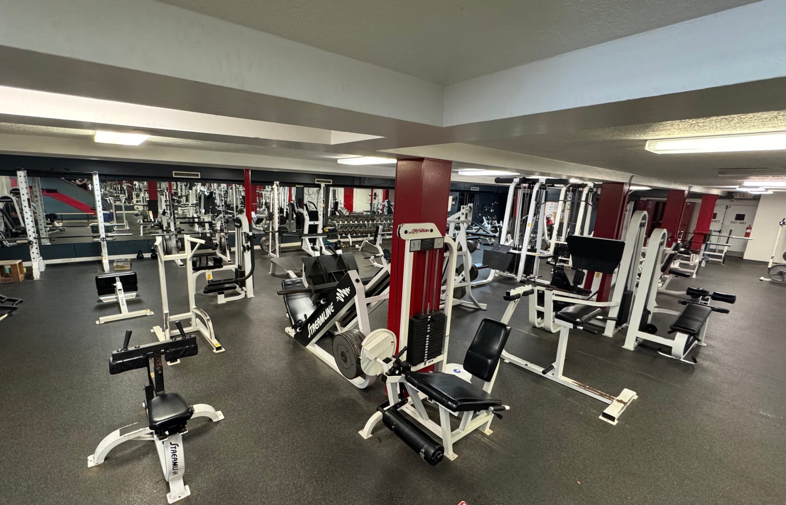 Empty gym with various exercise machines and weights, mirrors on the wall, and bright lighting.