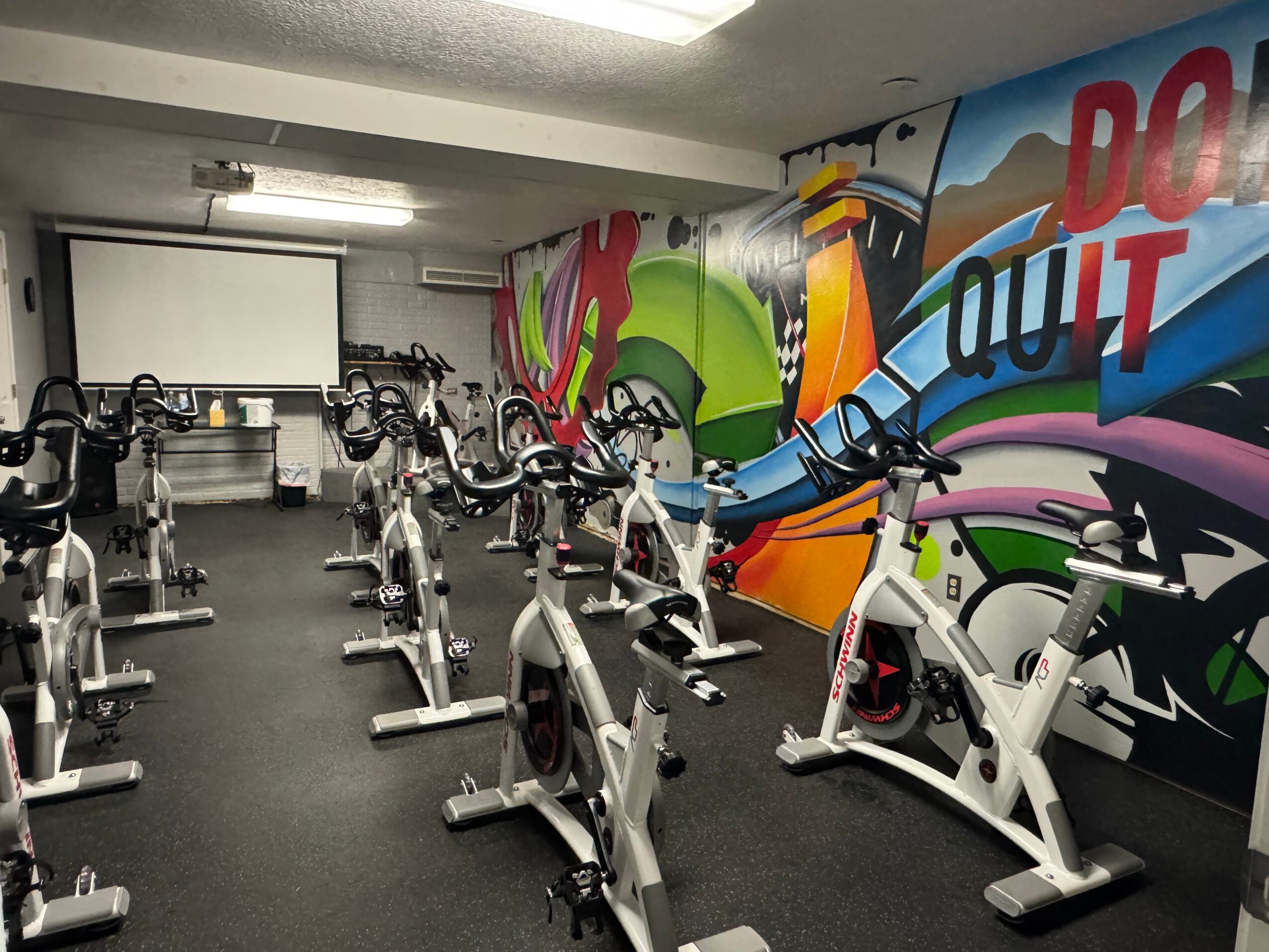 Indoor gym with stationary bikes and a colorful graffiti wall, ceiling lights, and a projector screen in the background.
