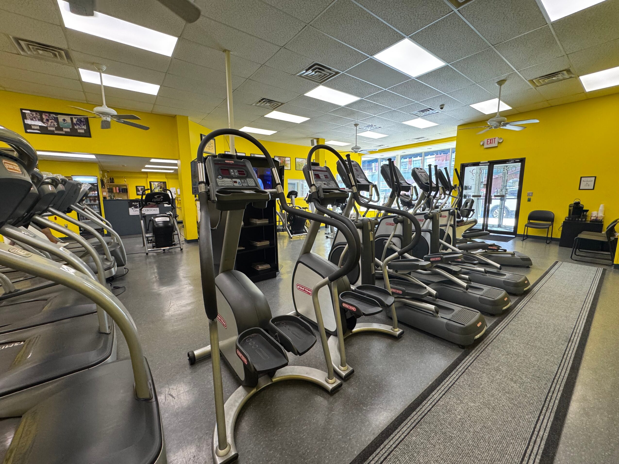 Gym with yellow walls, exercise machines, and treadmills in a row, bright lighting, and large windows.