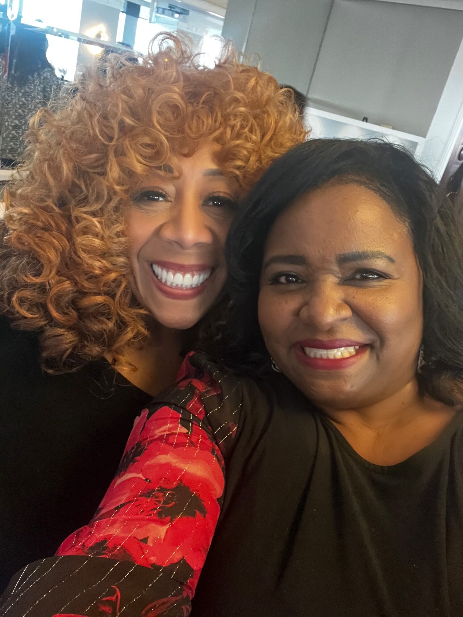 Two women smiling closely, one with curly red hair and the other with straight black hair, in an indoor setting.