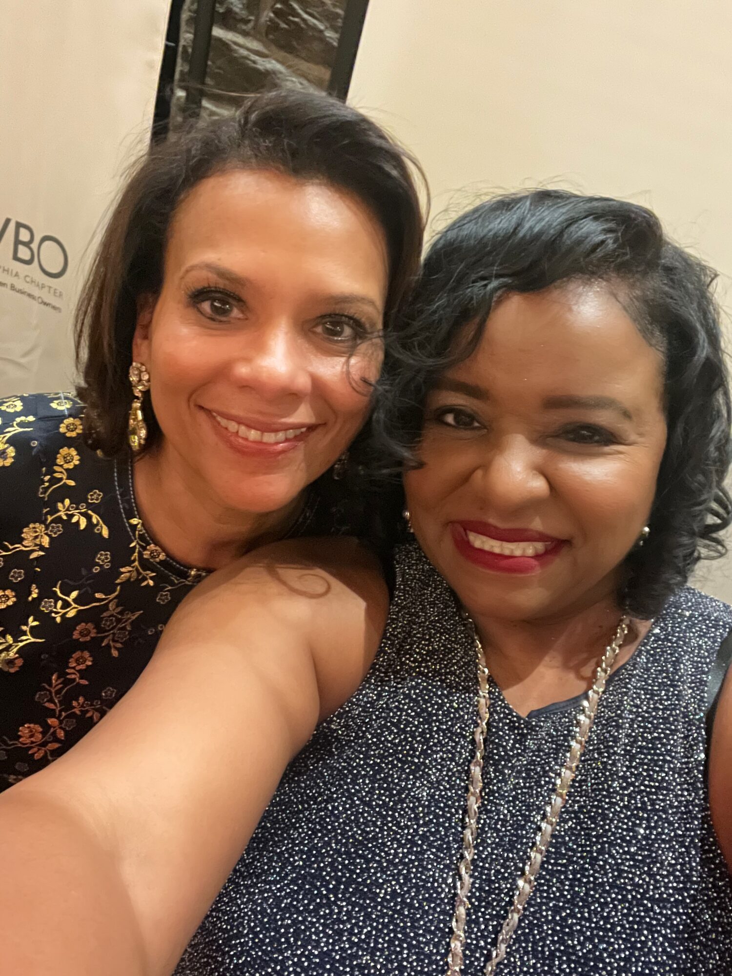Two women smiling closely for a selfie, one wearing a black and gold patterned top, the other in a dark, sparkly dress.