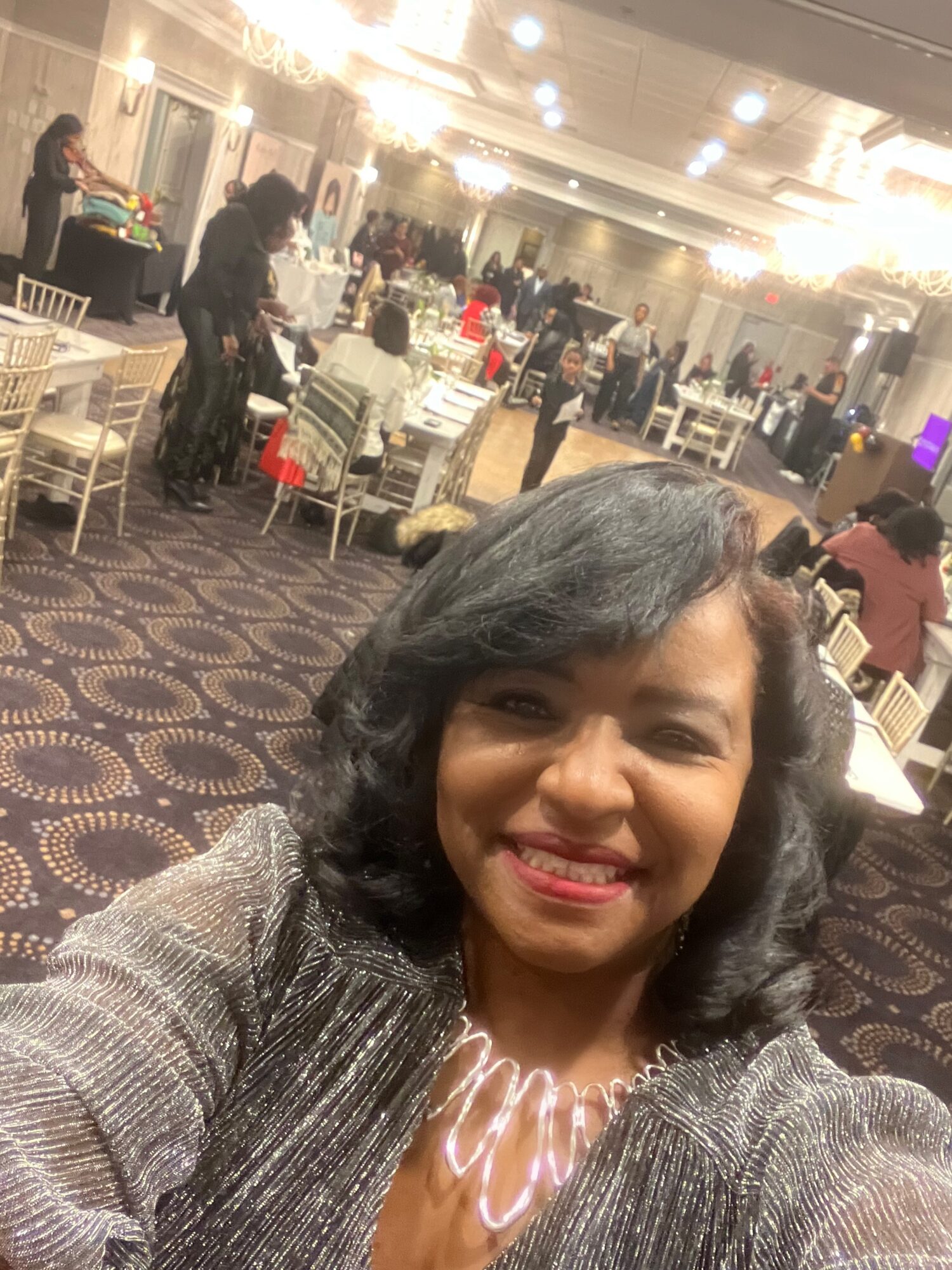 Smiling woman with shoulder-length dark hair in a shiny silver jacket at a banquet hall with tables and guests.