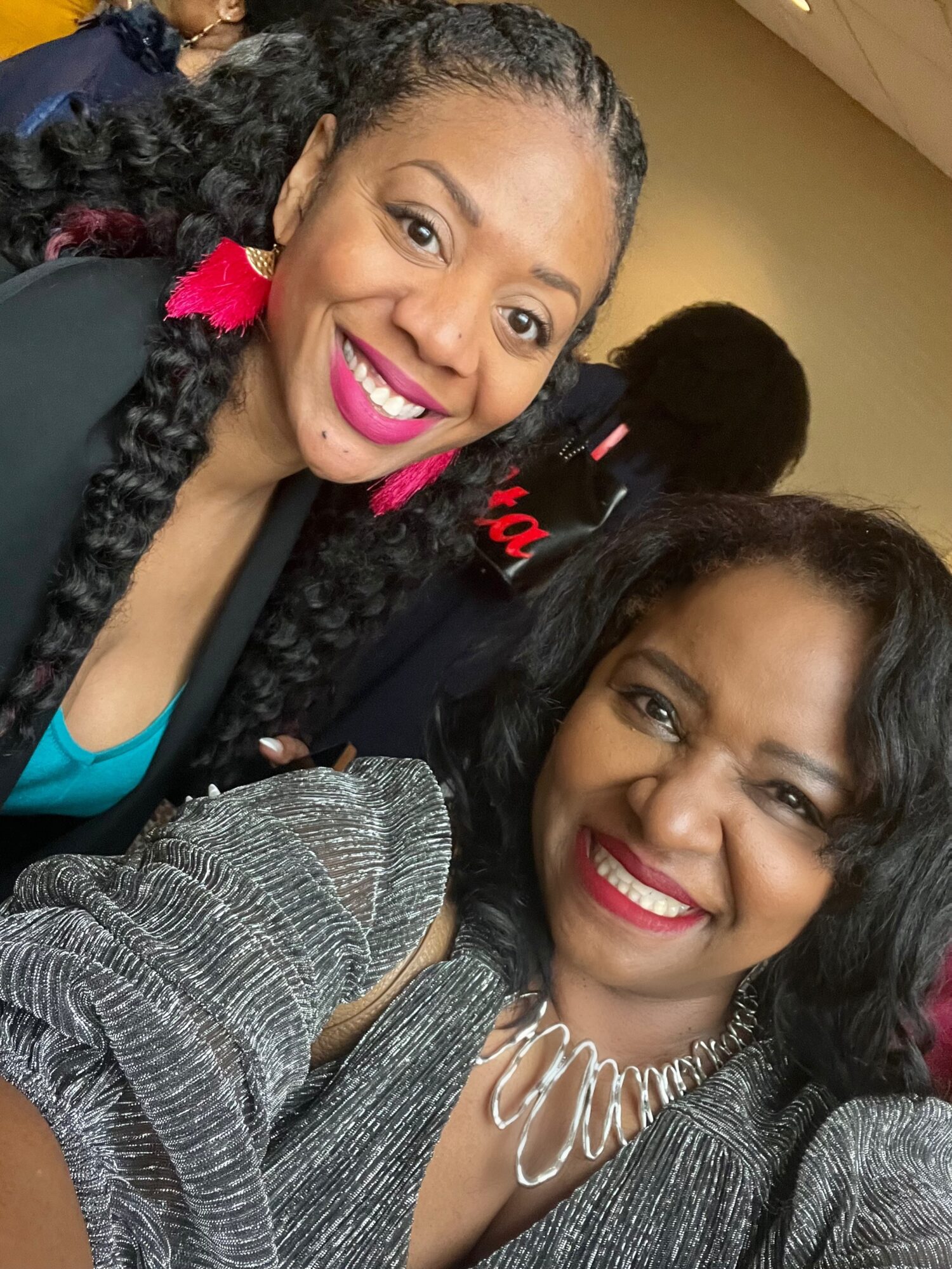 Two women smiling for a selfie, one with curly hair and bright lipstick, the other with wavy hair and a necklace.