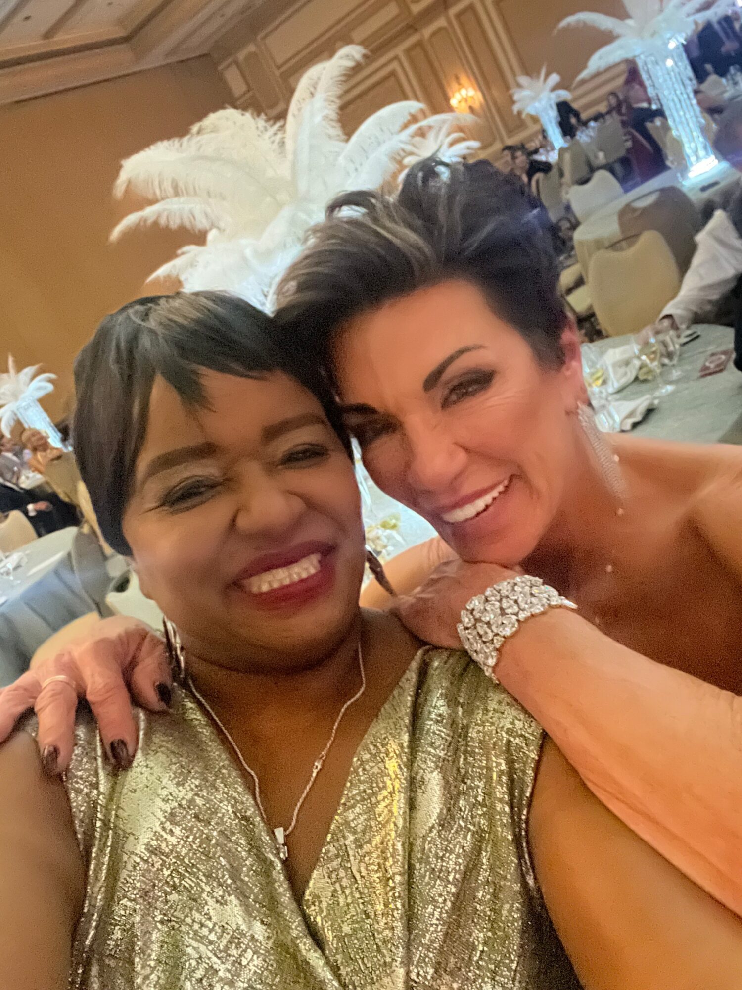 Two women smiling closely at a celebration with elegant decorations and feathered headpieces in the background.