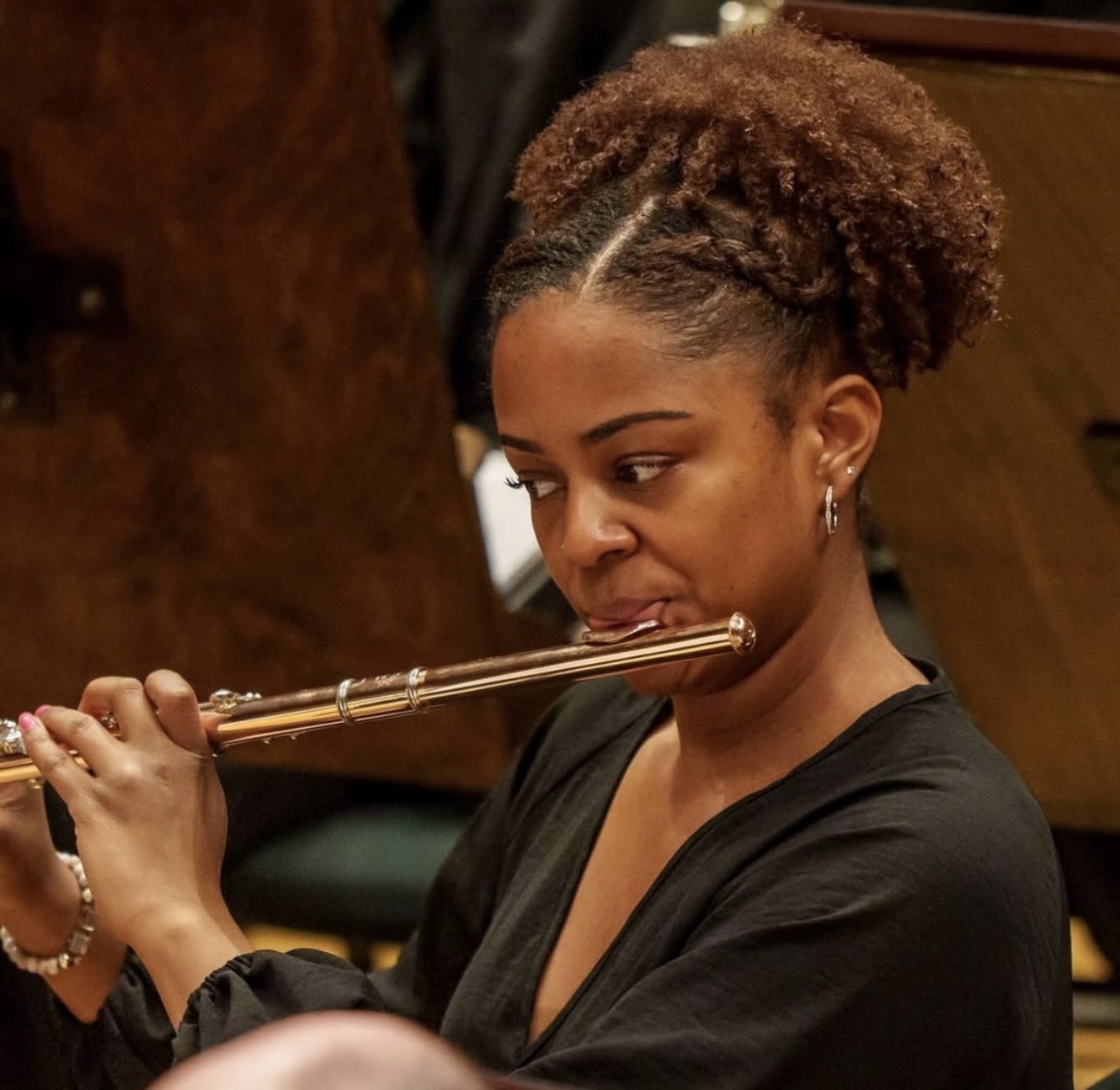 Woman playing flute, focused, with curly hair styled in an updo, wearing a black top, in a music setting.
