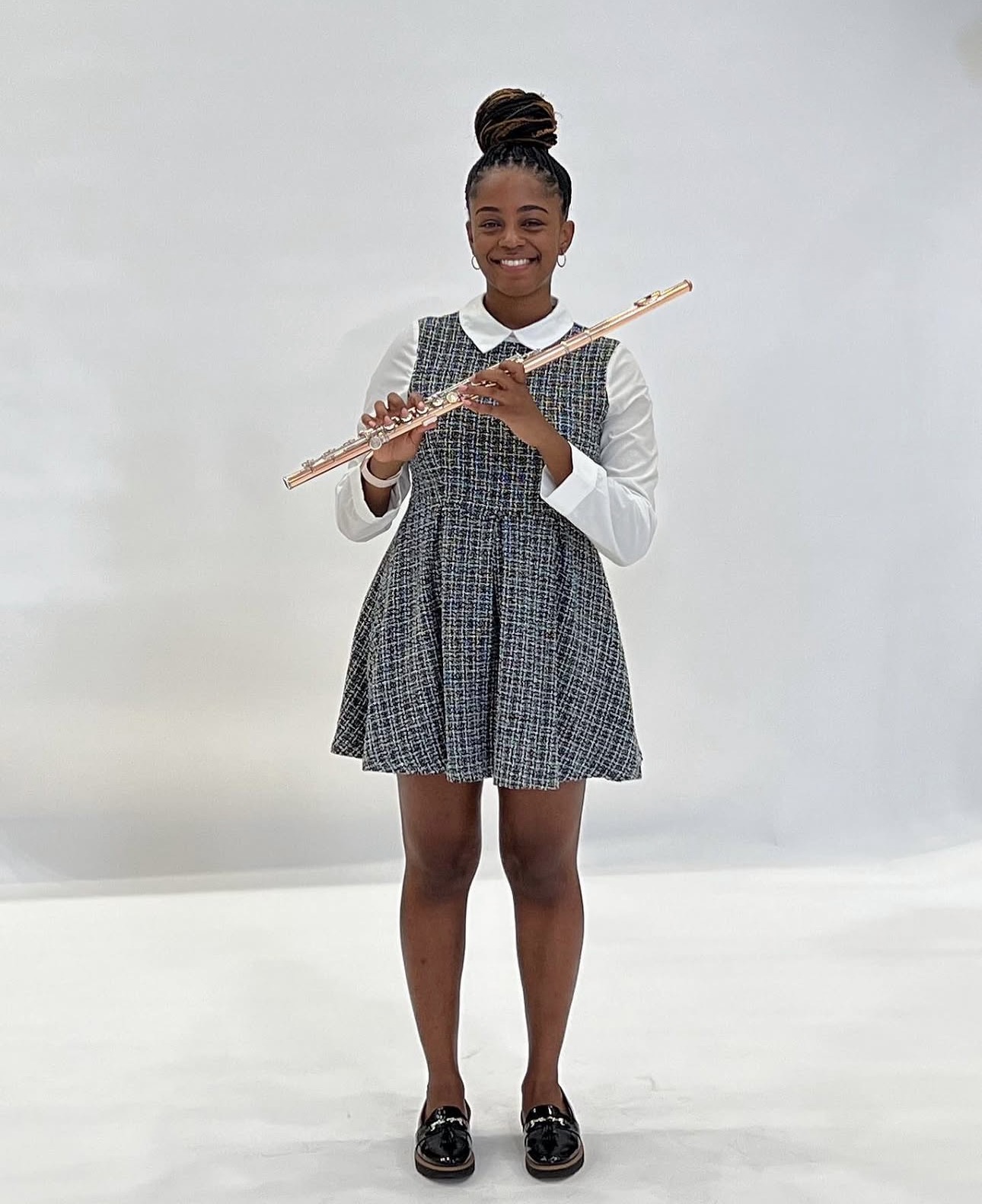 Young girl smiling, holding a flute, wearing a school uniform with a plaid dress and white shirt, standing against a plain background.