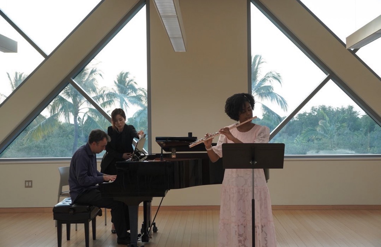 Three musicians perform in a room with large windows showing palm trees outside, one playing piano, one on violin, one on flute.