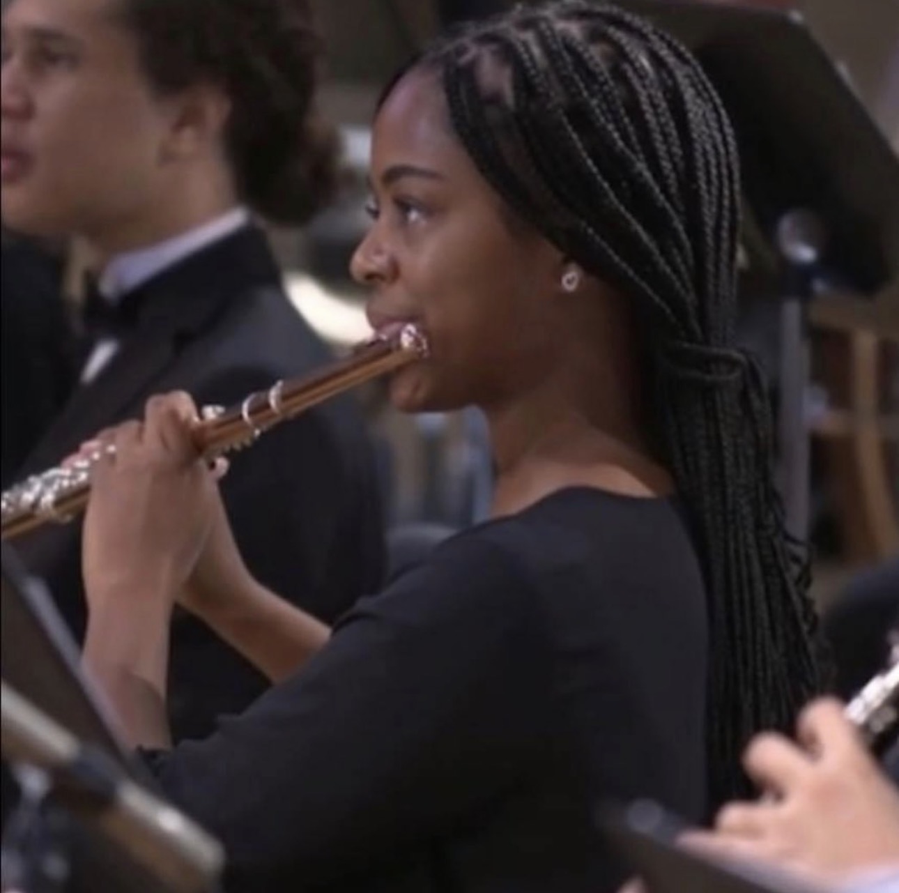 Young woman playing flute in an orchestra, with other musicians in the background, focused on her performance.