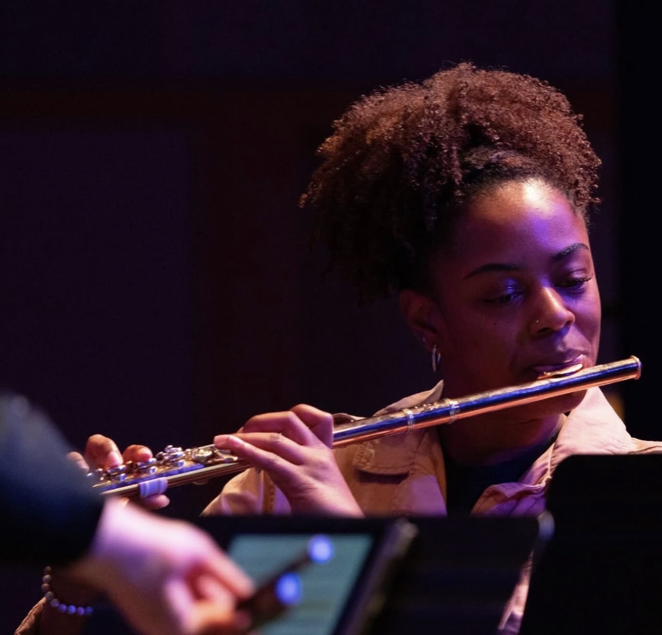 Woman playing flute, focused, with dark background, curly hair, and beige clothing.