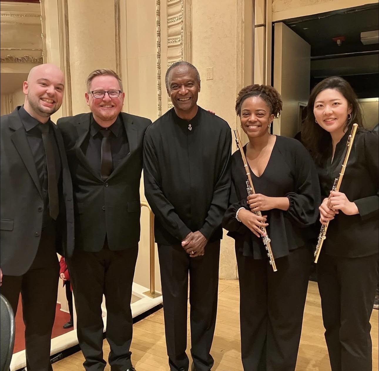 Five people in formal attire standing indoors, two holding flutes, smiling at the camera.