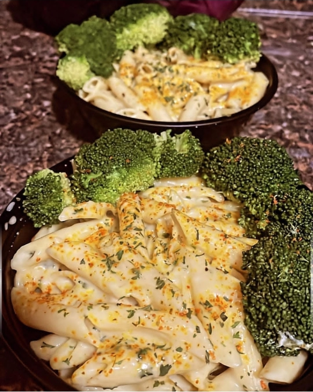 Two bowls of pasta with broccoli, garnished with herbs and cheese, on a dark surface.