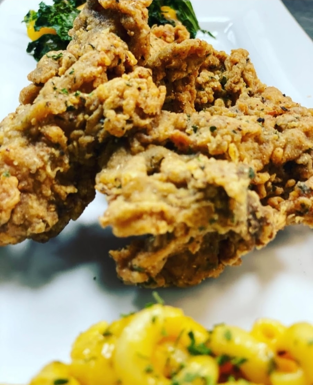 Close-up of fried chicken pieces on a white plate with some greens in the background, and a side of yellow pasta salad with herbs.