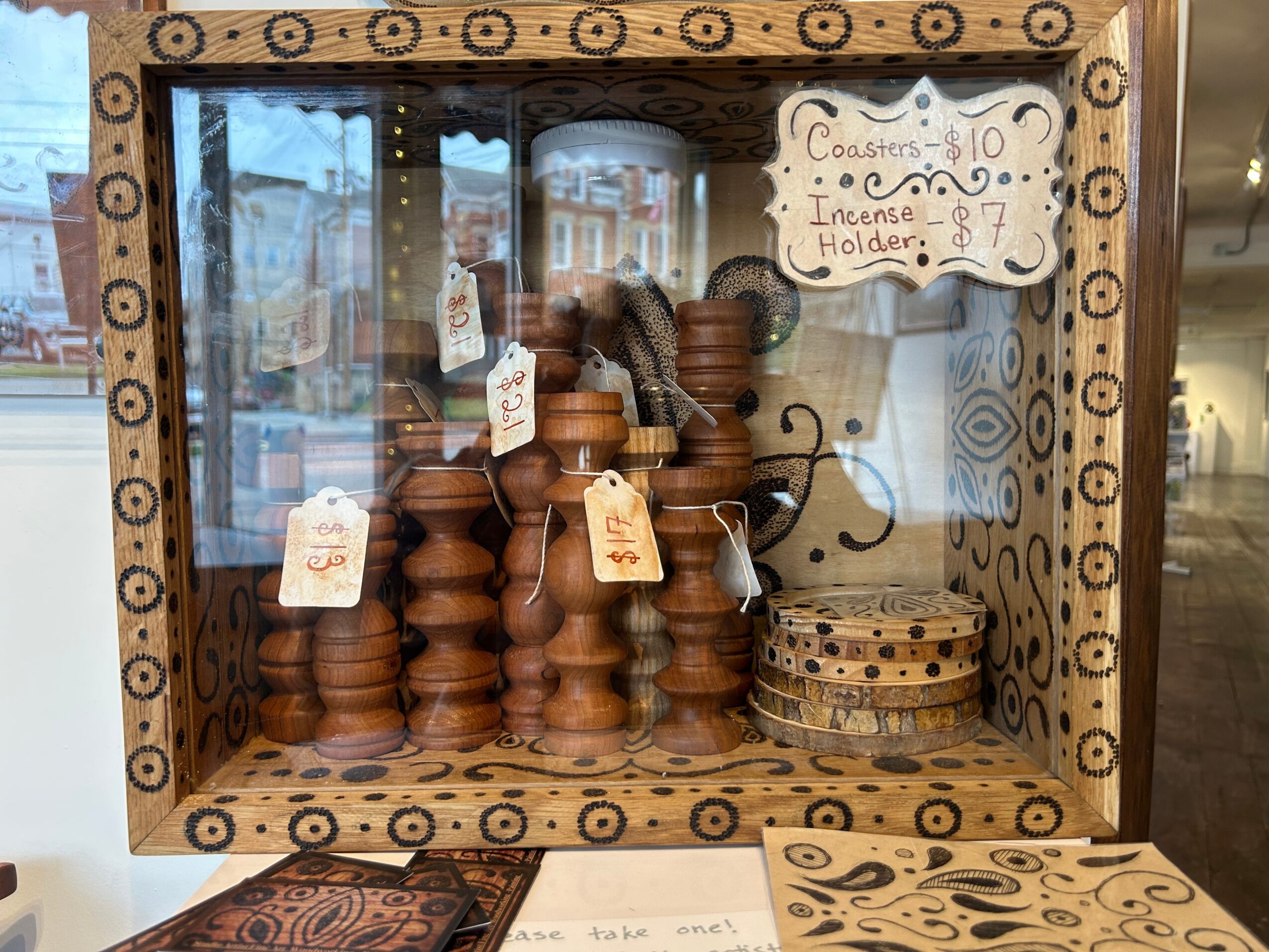 Wooden display case with incense holders and coasters, handwritten signs, and a reflection of a window in the background.