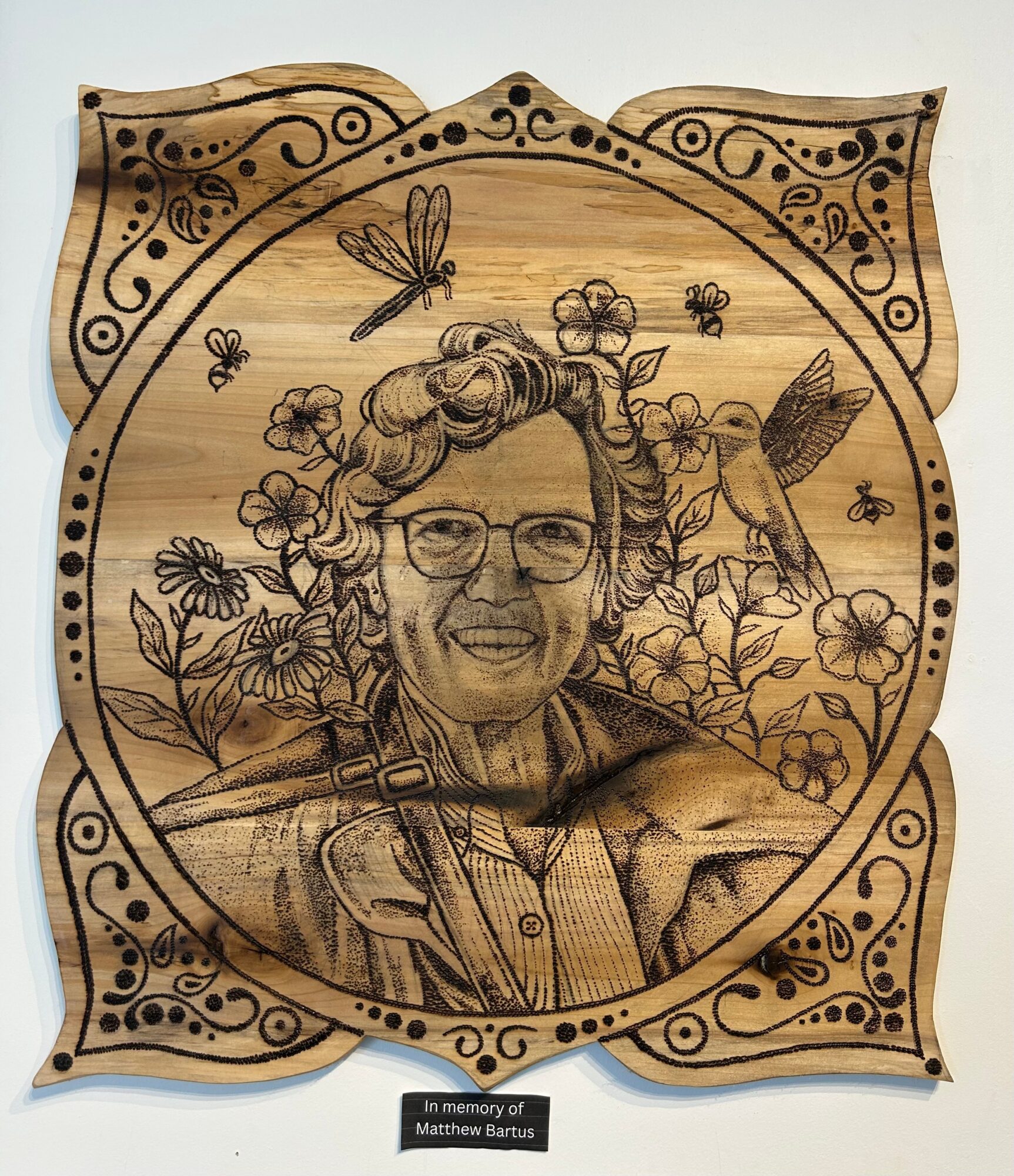 Wooden plaque with engraved portrait of a woman wearing glasses, surrounded by flowers, insects, and a bird, with decorative border.