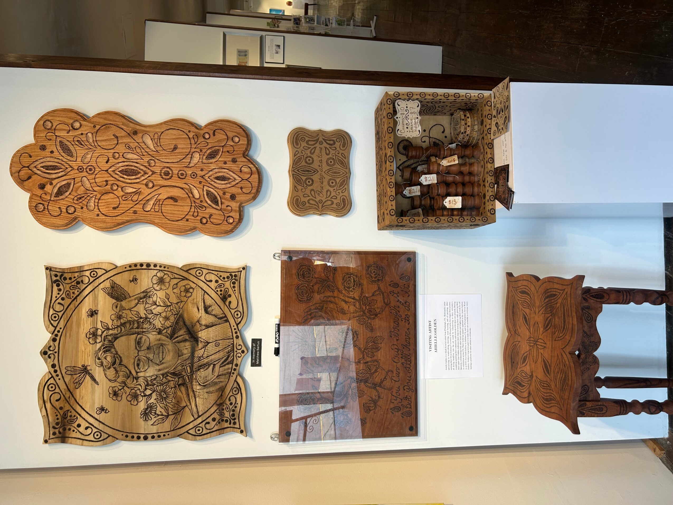 Display of wooden artifacts including carved panels, a basket, and a small table, arranged on a white wall and surface.