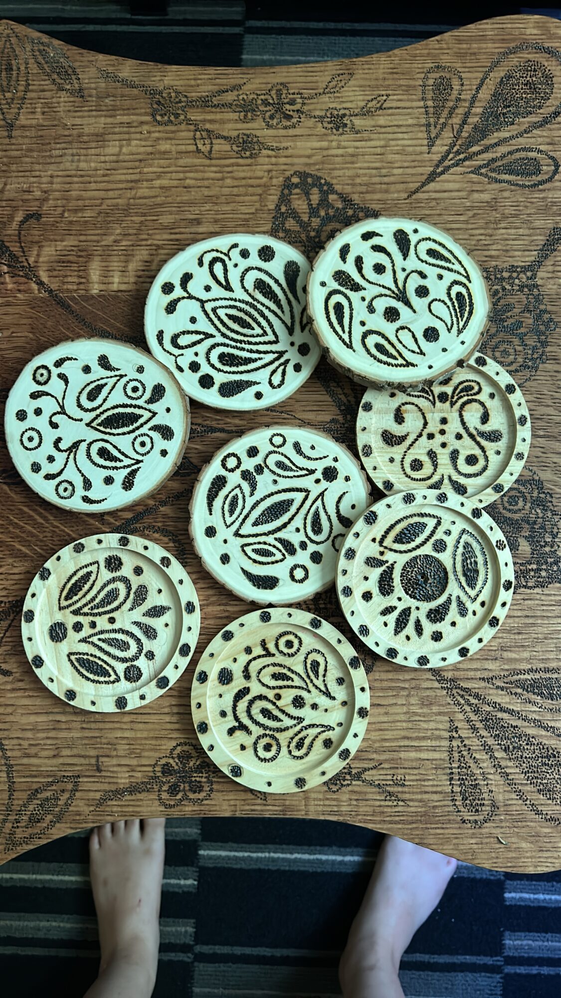 Eight round, patterned objects with intricate designs, placed on a wooden surface with floral and leaf patterns, two feet visible at bottom.