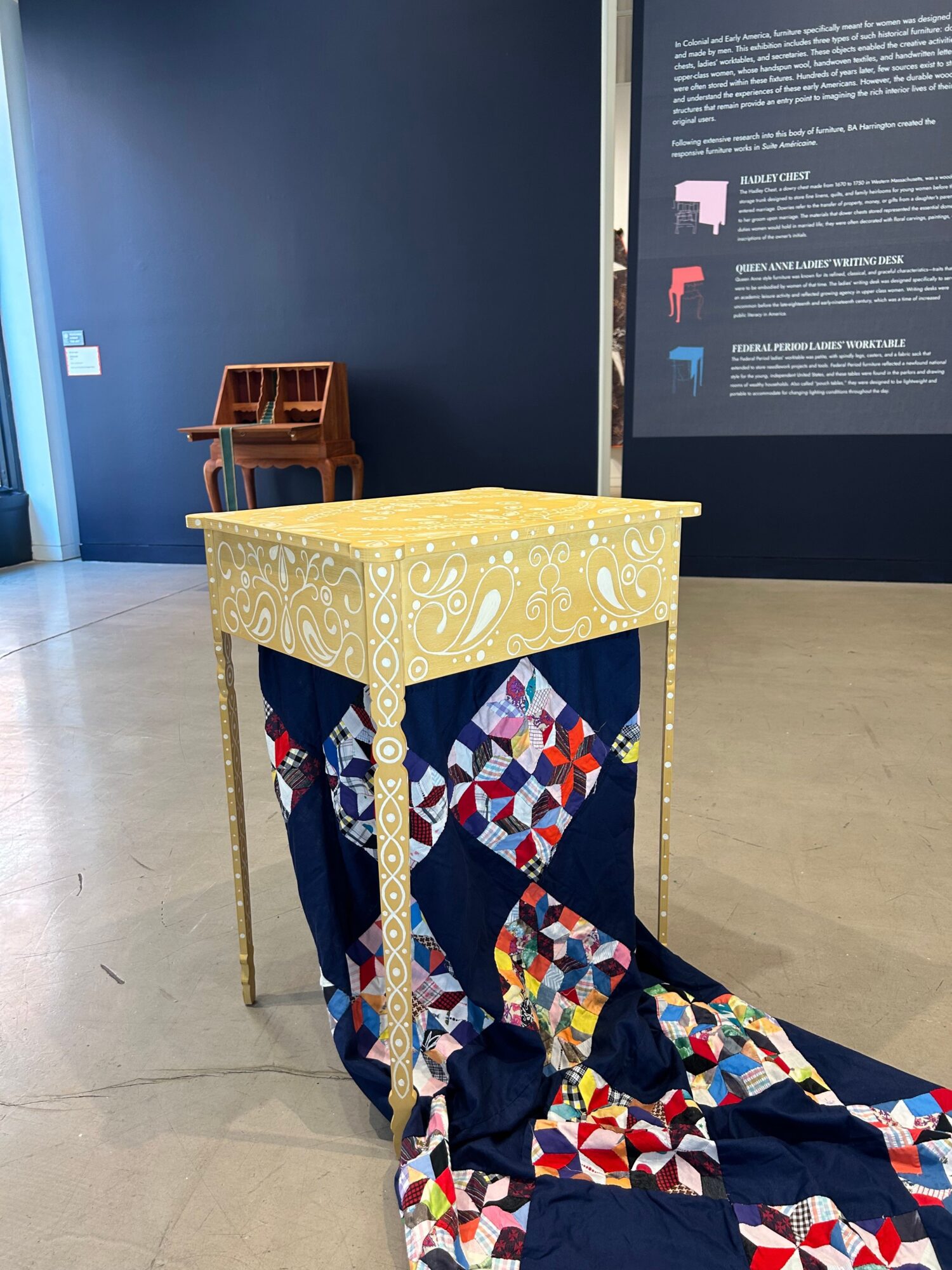 Table with a yellow tablecloth, colorful quilt draped over the side, and a dark blue wall with a display screen in the background.