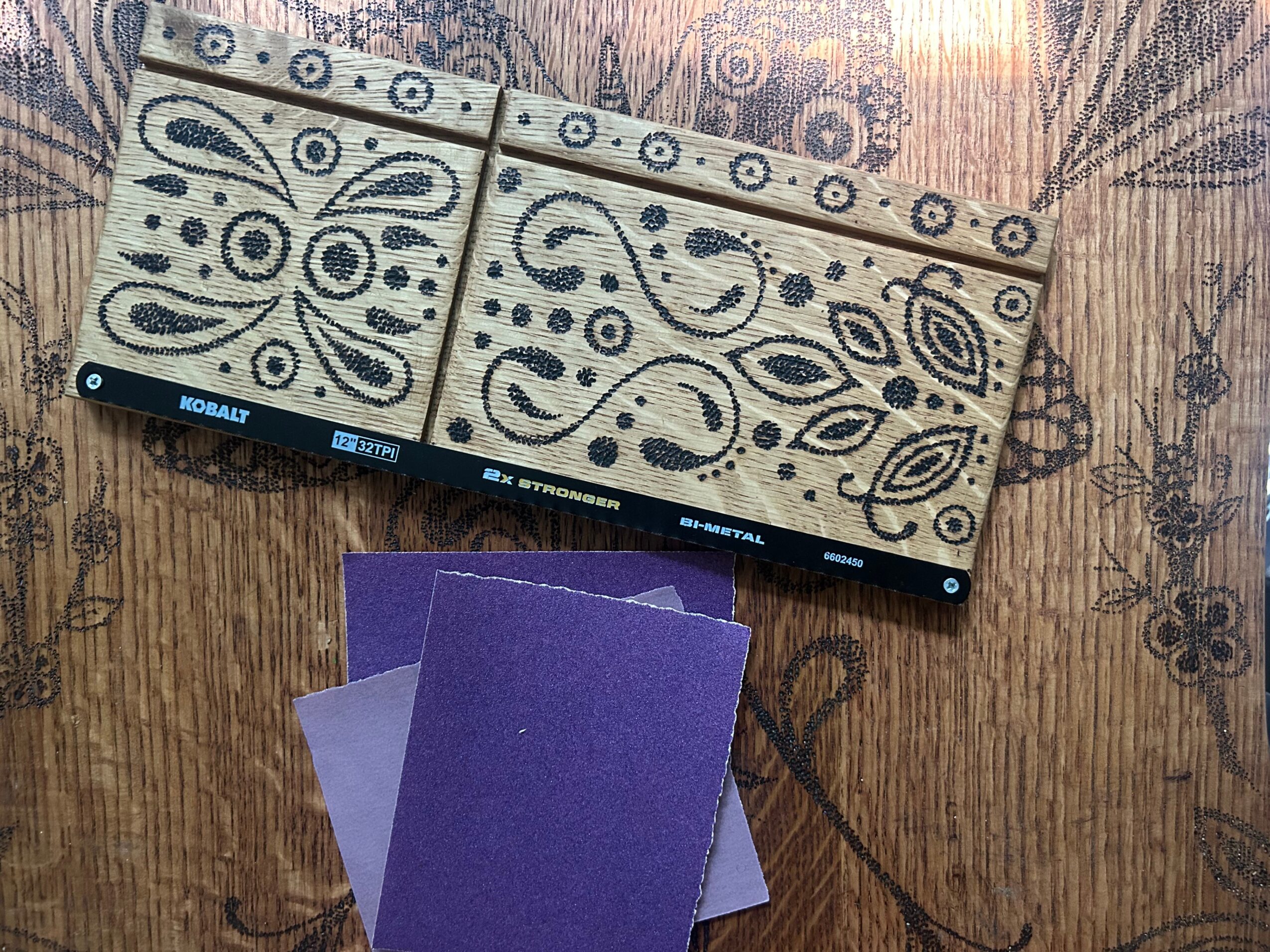 Box of wooden dominoes with black patterns, two purple domino tiles, on a wooden surface with floral design.