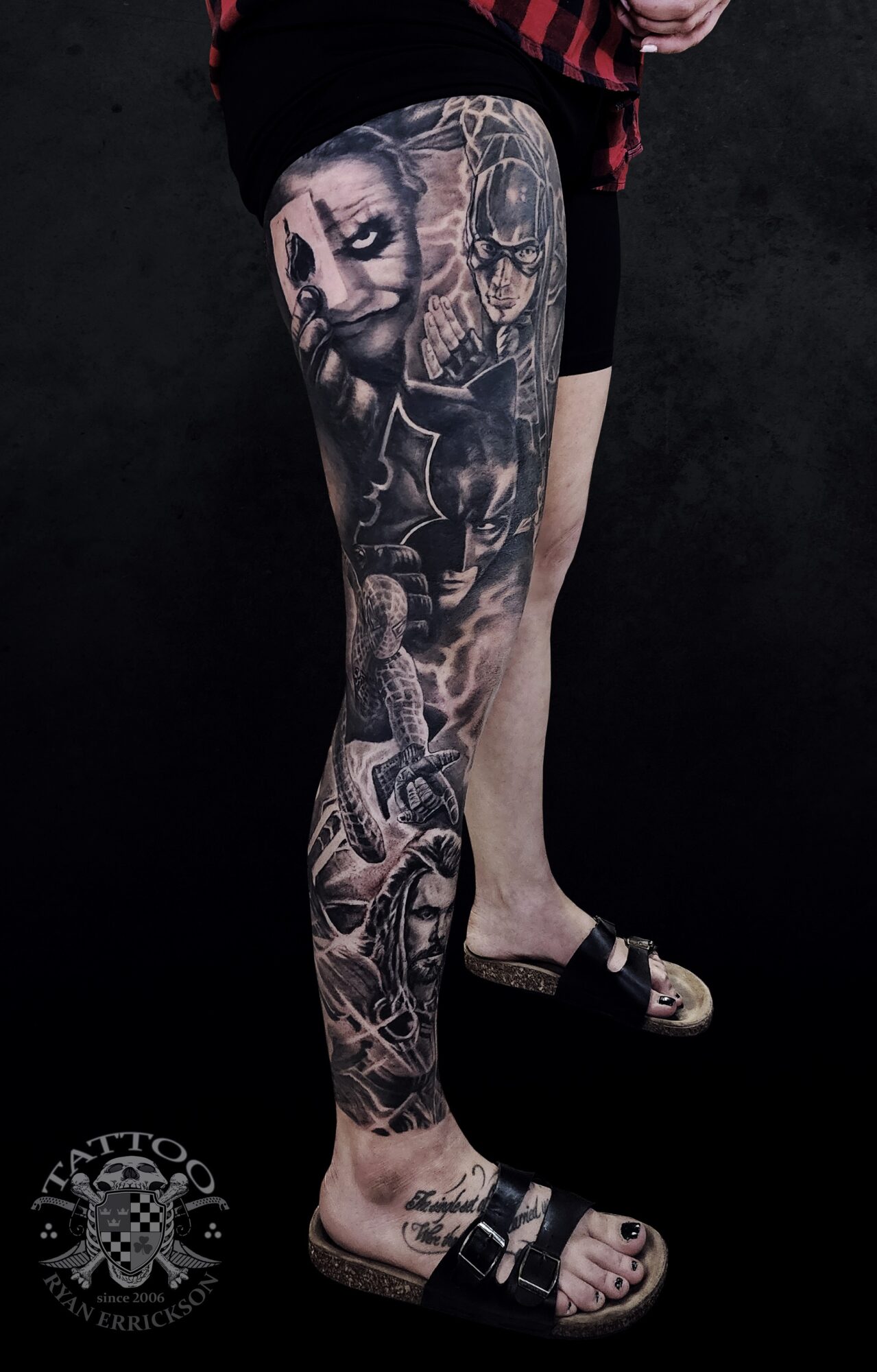 Person's leg with detailed black and gray tattoo featuring faces, skulls, and abstract designs, against a dark background.
