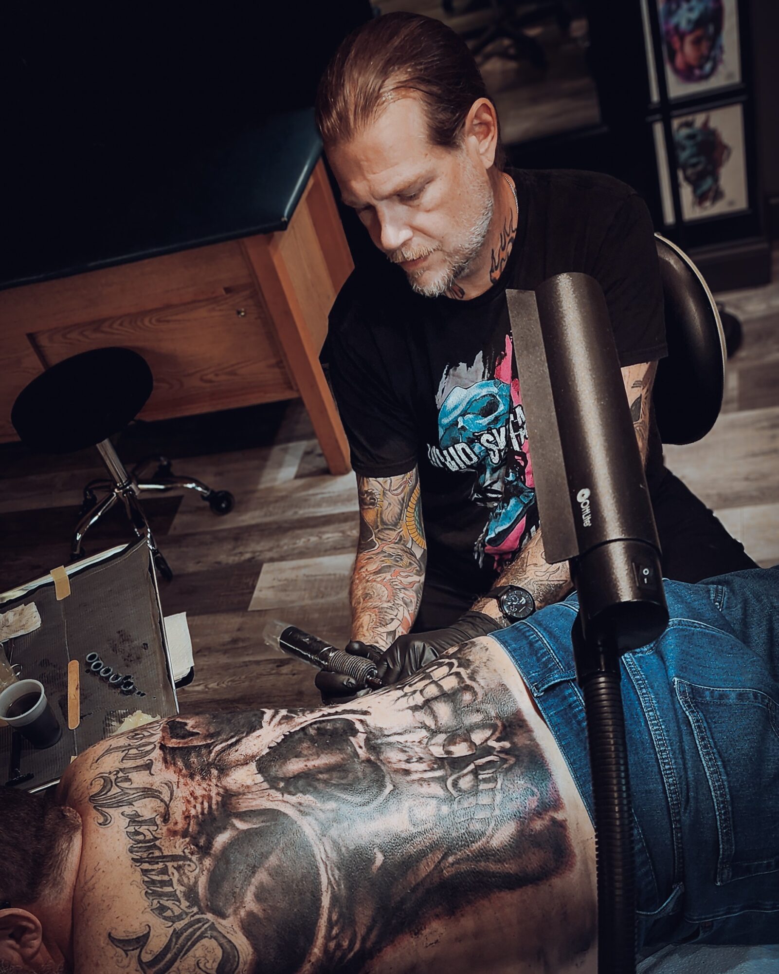 Tattooed man lying on his stomach, receiving a tattoo from a tattoo artist in a studio.