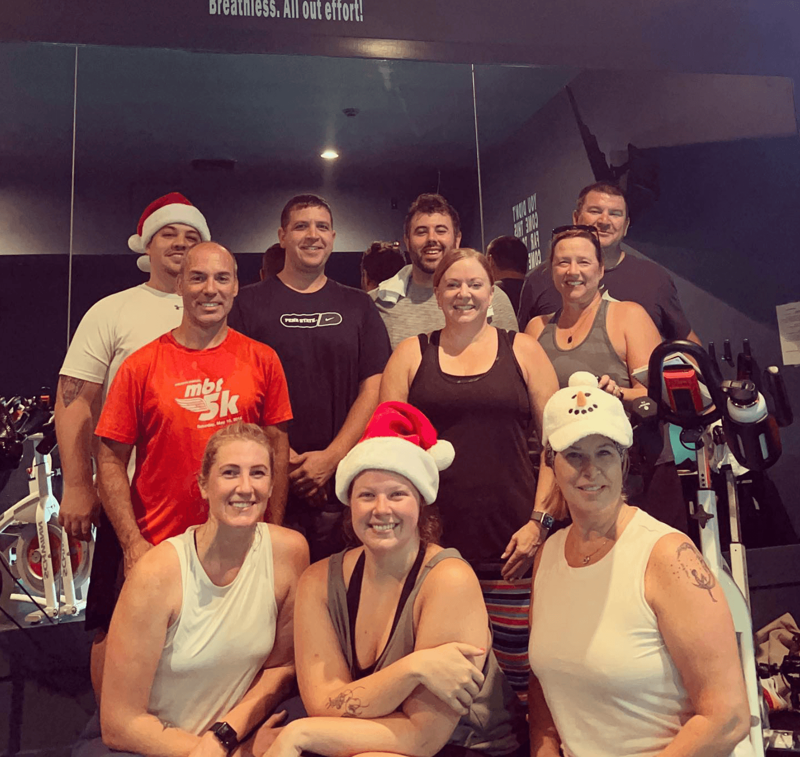 Group of nine people in a gym, some wearing Santa hats, smiling, with gym equipment in the background.