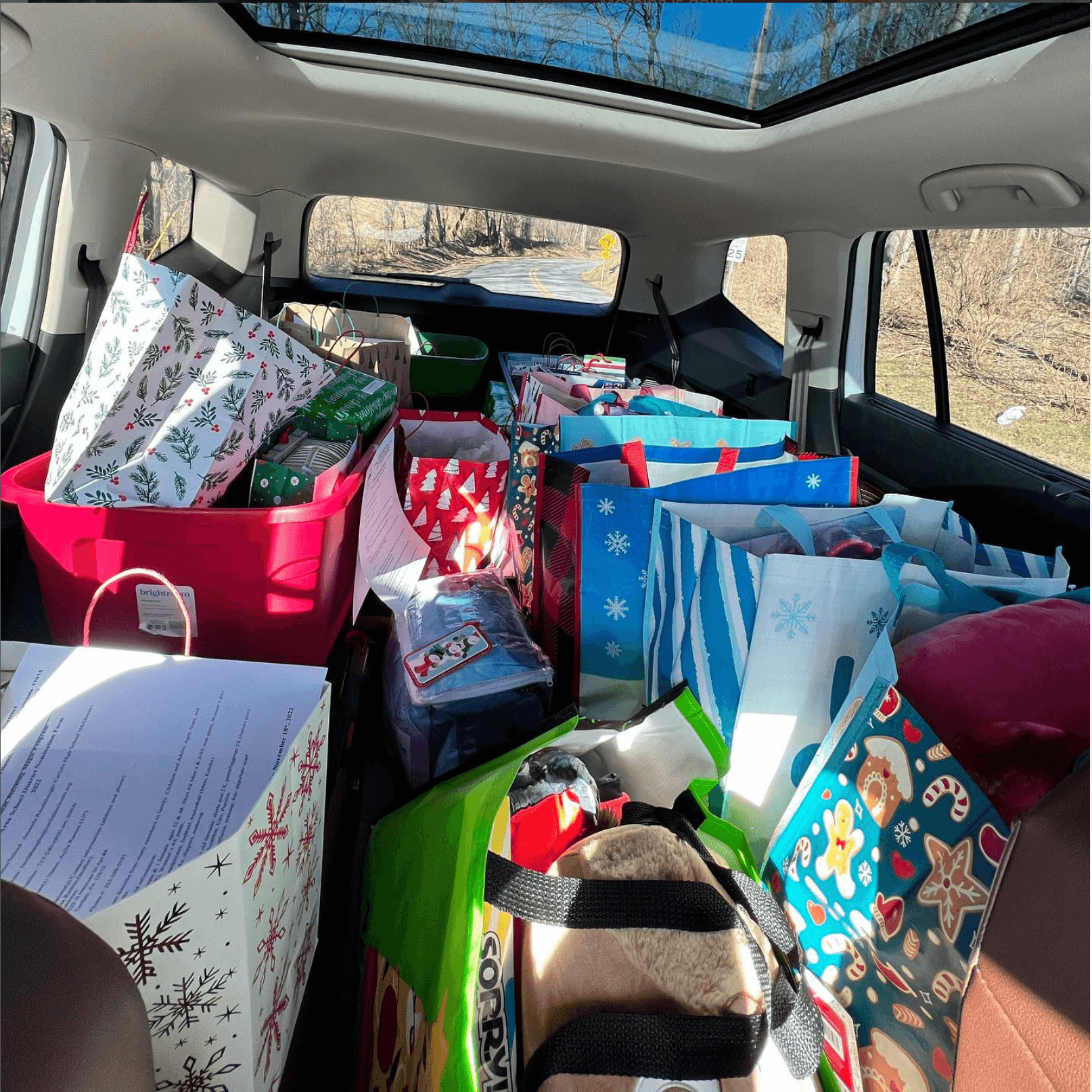 Car interior filled with wrapped presents and gift bags, sunlight through sunroof, outdoor scenery visible through windows.