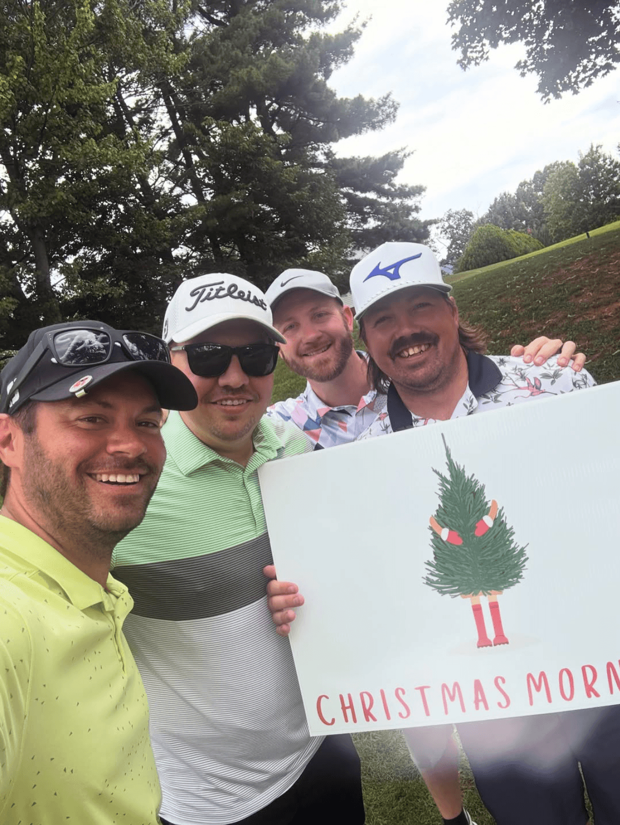 Four men smiling outdoors, holding a Christmas sign with a tree and text, wearing golf caps and sunglasses.