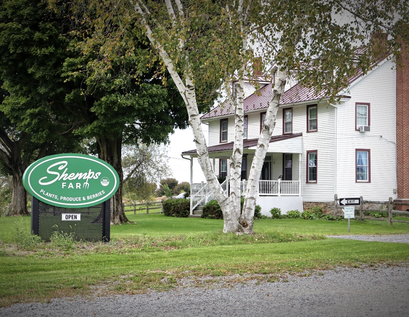 Farm stand sign for Shemp's Farm with a white house and trees in the background, green grass in front.