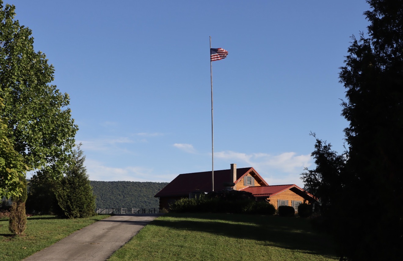 A building with a red roof and an American flag on a tall flagpole in front, surrounded by trees and a clear blue sky.