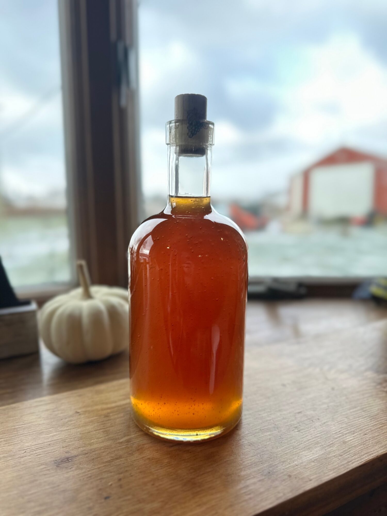 Glass bottle with amber liquid on wooden surface, window and sky in background.